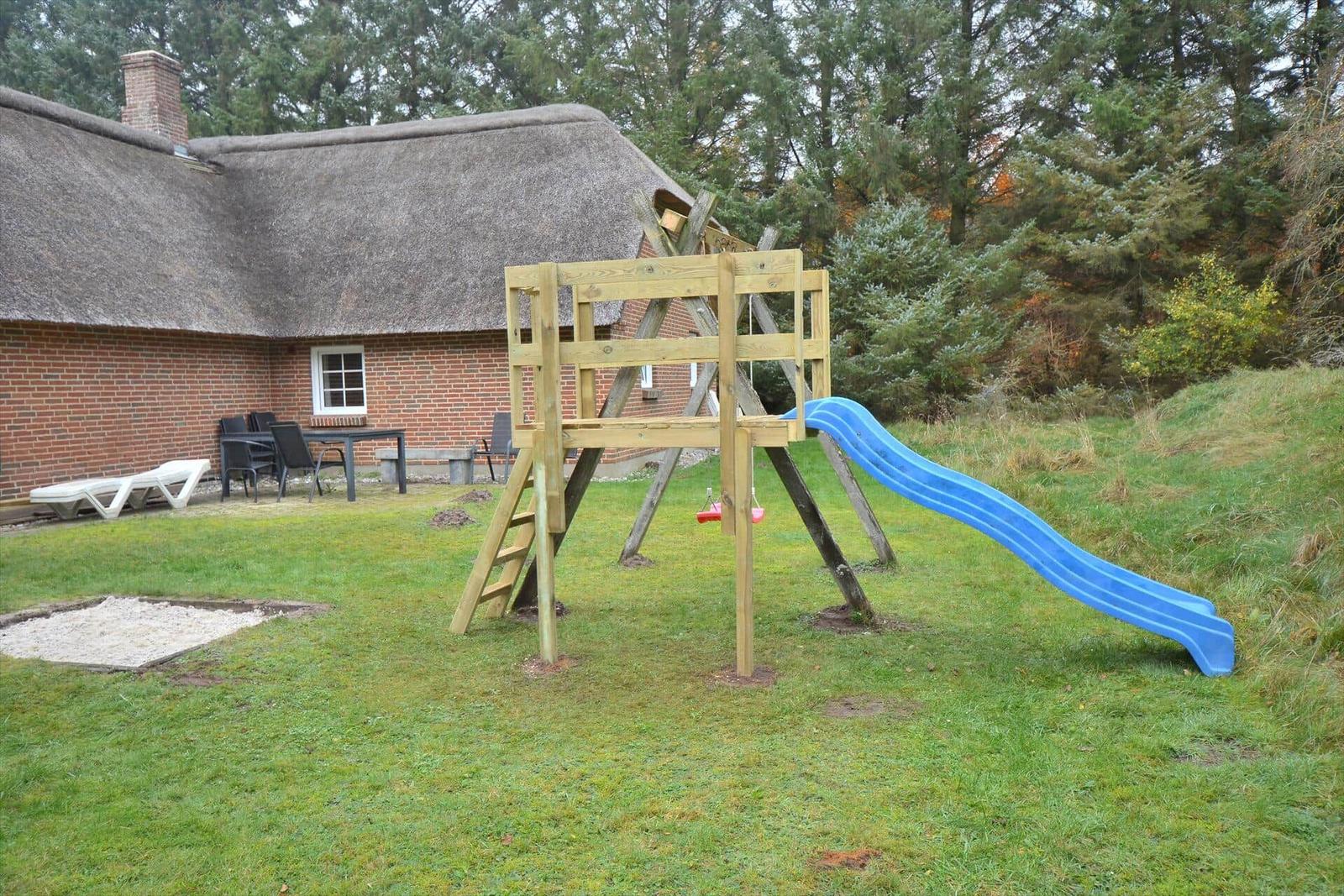 Hinterhof mit Holzspielplatz, blauer Rutsche und Sandkasten. Im Hintergrund ein Haus mit Strohdach.