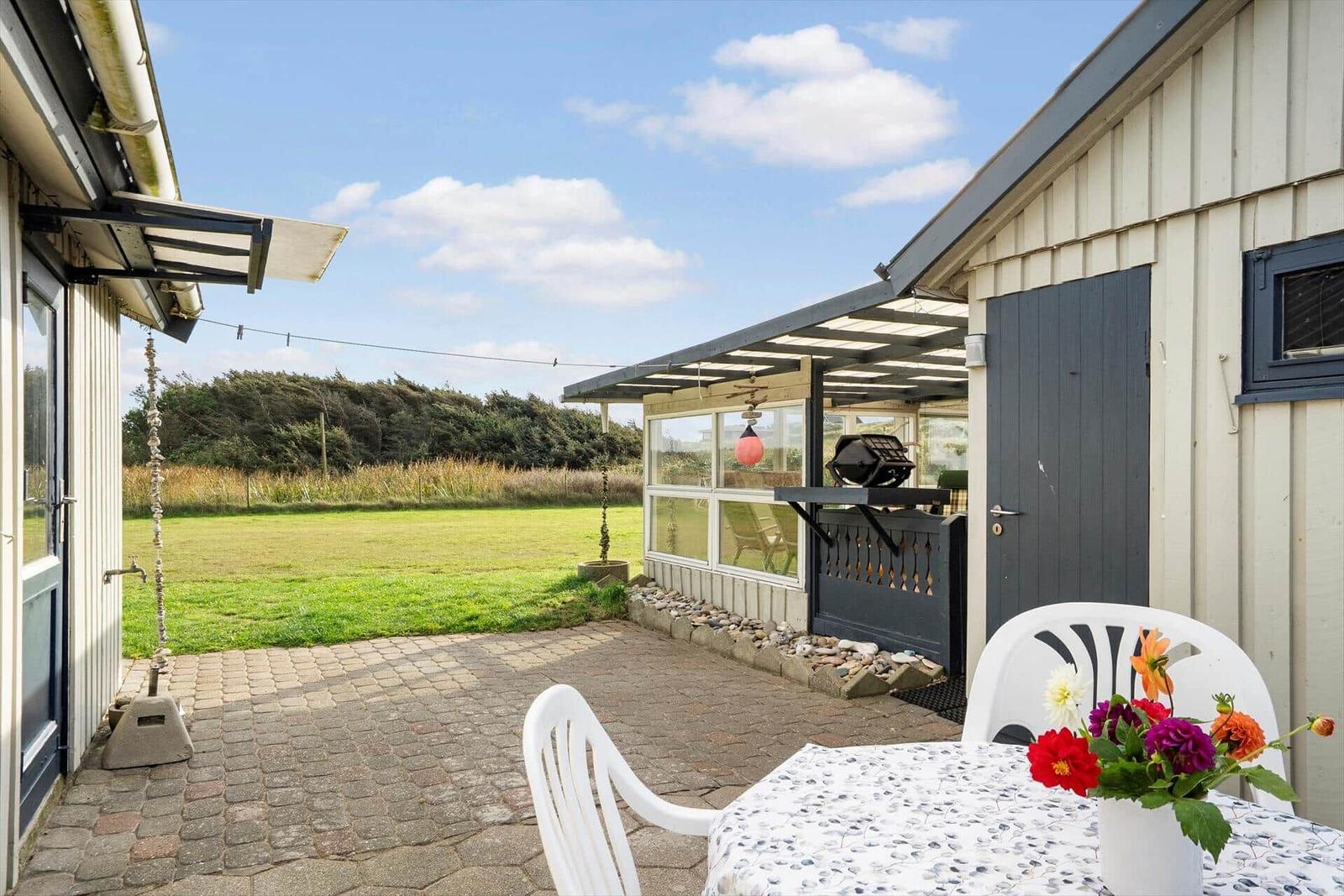 Terrasse mit Tisch, Stühlen und Blumen. Hintergrund: Grasland und Bäume.