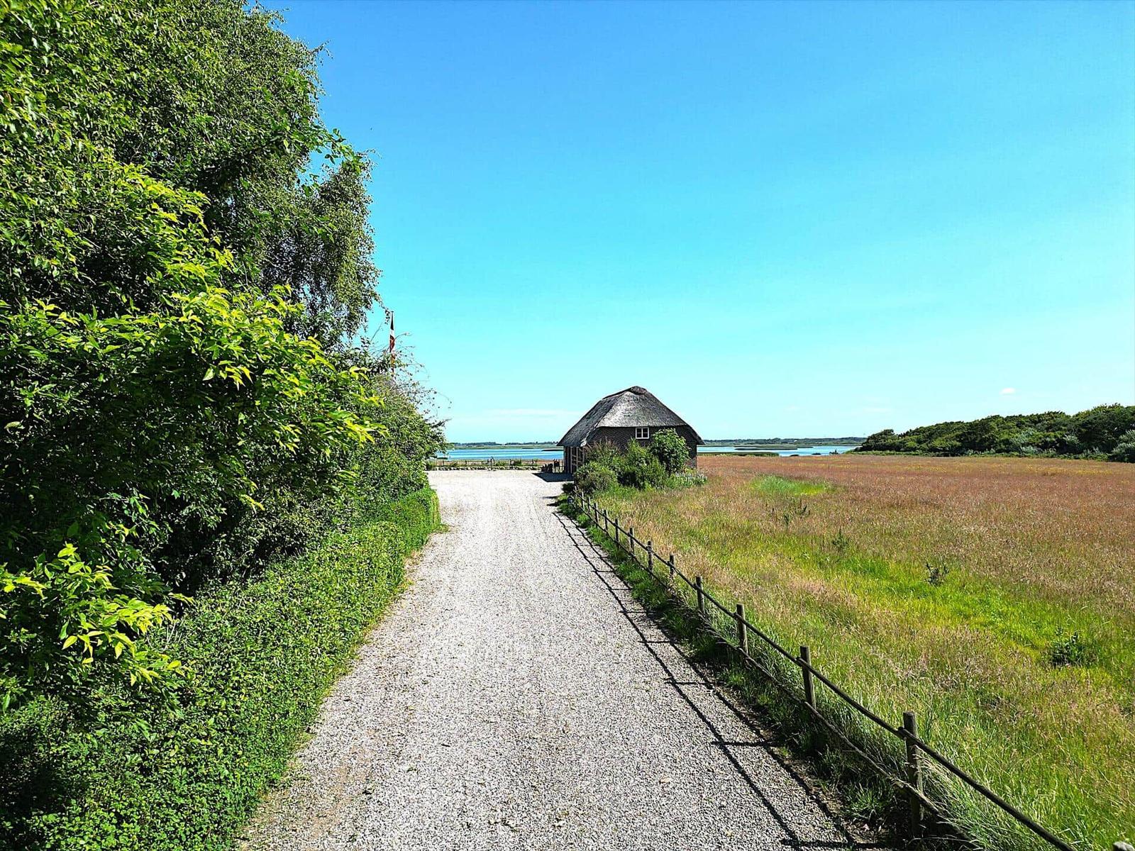 Hütte am Wasser mit Grünstreifen und Zaun entlang eines Kieswegs.