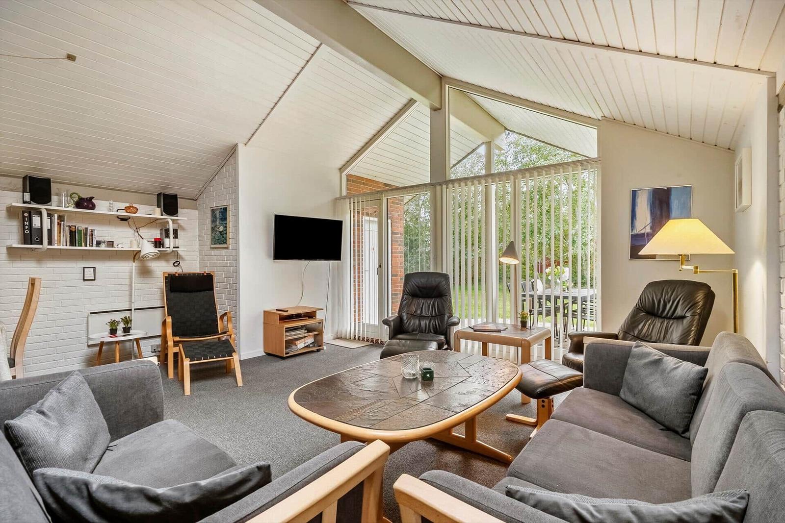 Wohnzimmer mit Sofa, Sessel und TV. Große Glasfront mit Blick auf Garten.
