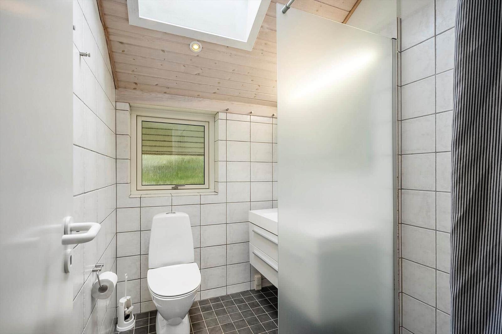 Badezimmer mit Toilette, Dusche und Fenster. Holzdecke und Fliesenwand.