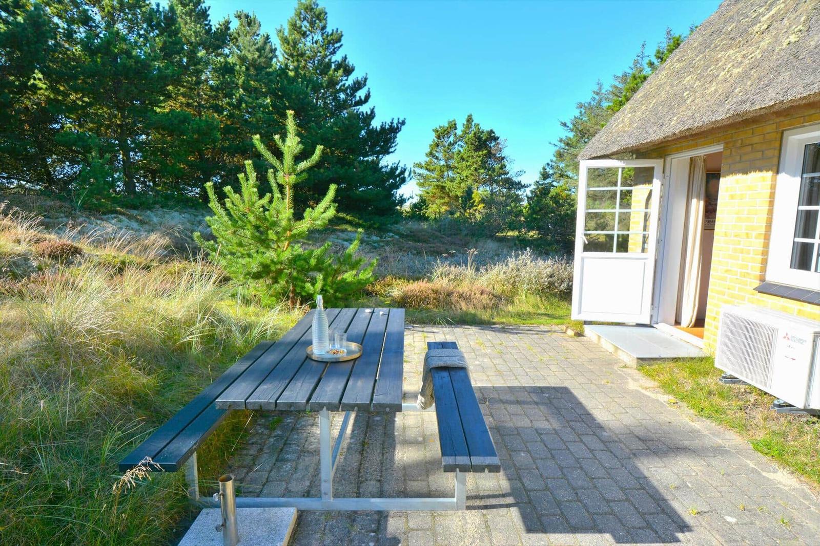 Hütte mit Holzterrasse, Tisch und Bänken im Wald. Tür offen, Blick auf Gras und Bäume.
