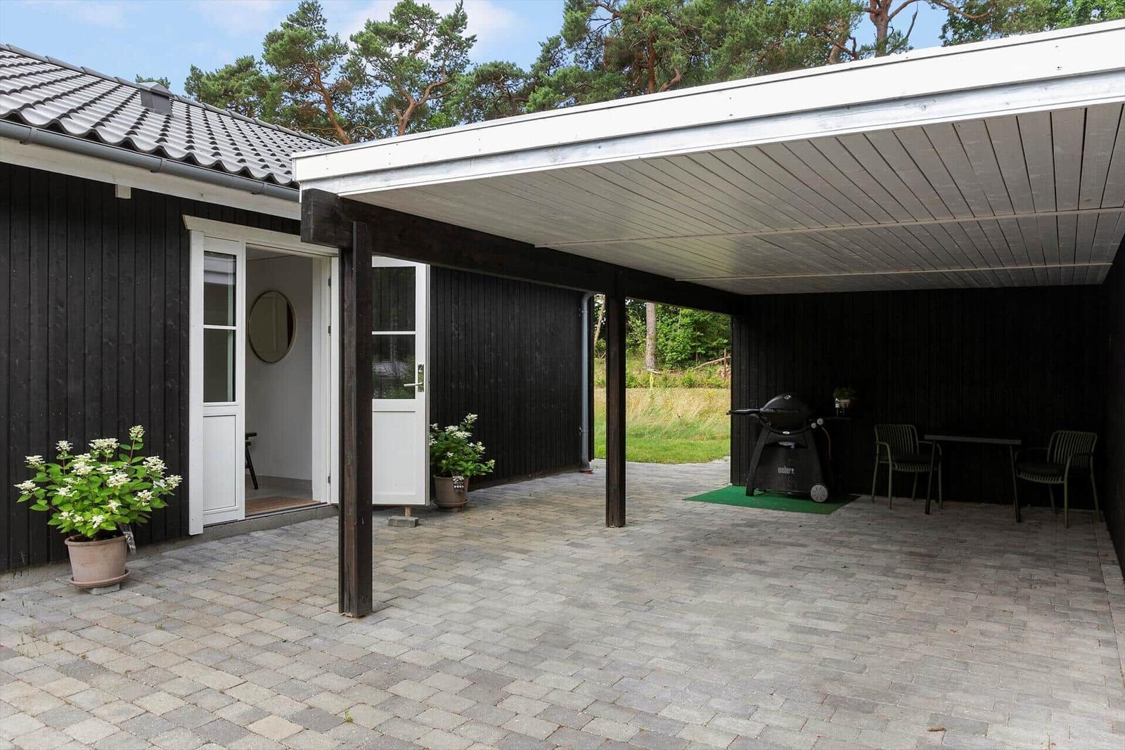 Terrasse mit Grill, Tisch und Stühlen unter Dach. Schwarze Holzwand, Pflanzen und Gartentür.