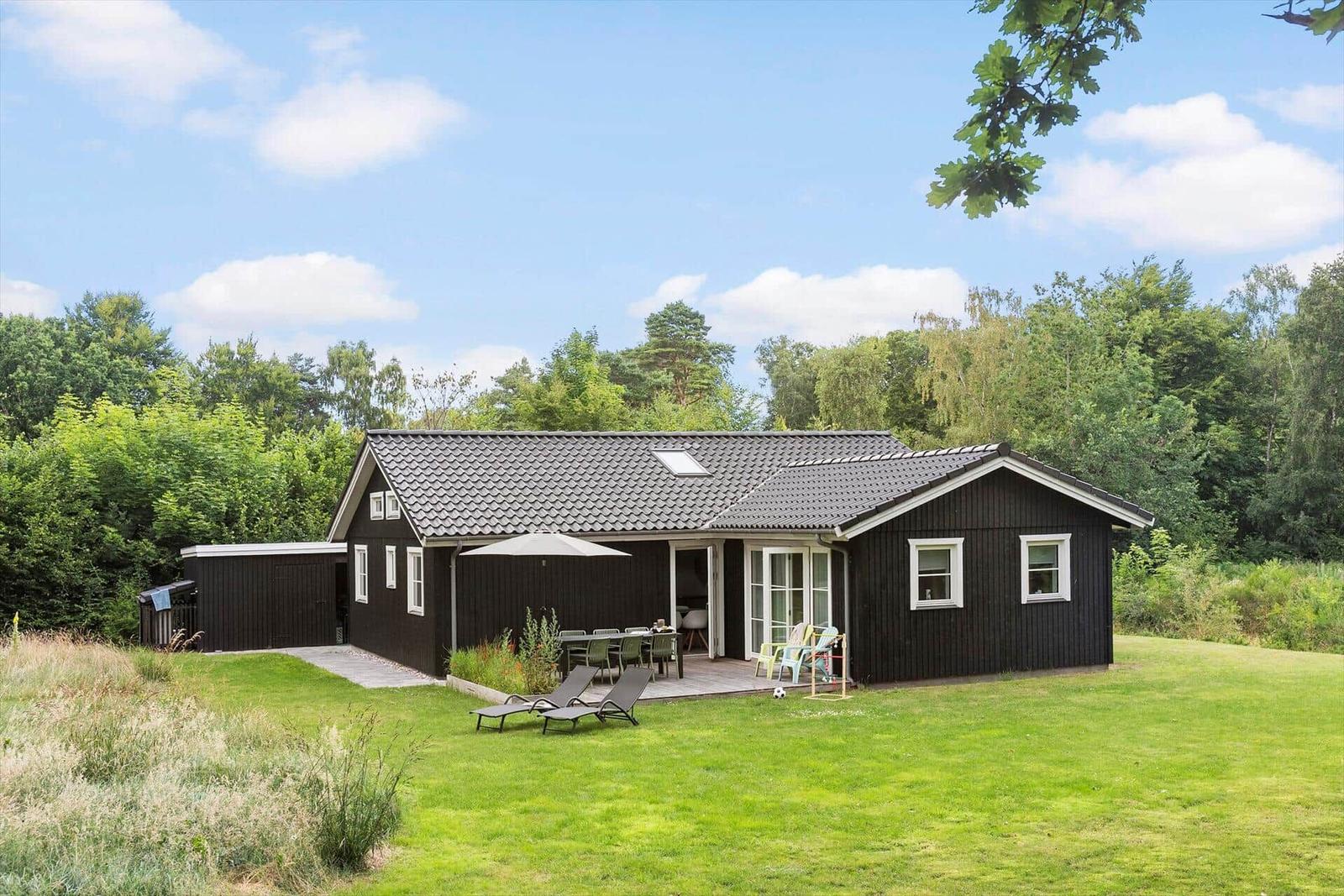 Schwarzes Holzhaus mit Terrasse, Garten und Grillplatz. Umgeben von Wald und Grünflächen.