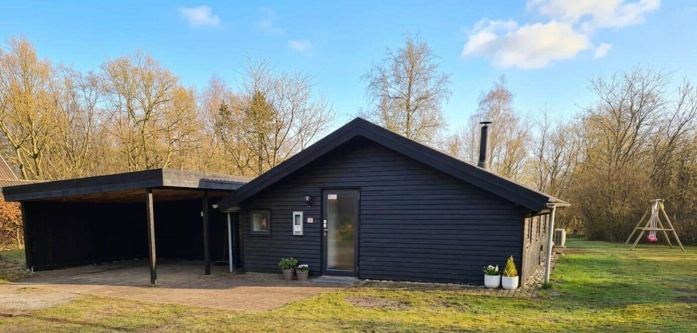 Schwarzes Holzhaus mit Carport und Garten. Hintergrund: Bäume und klarer Himmel.