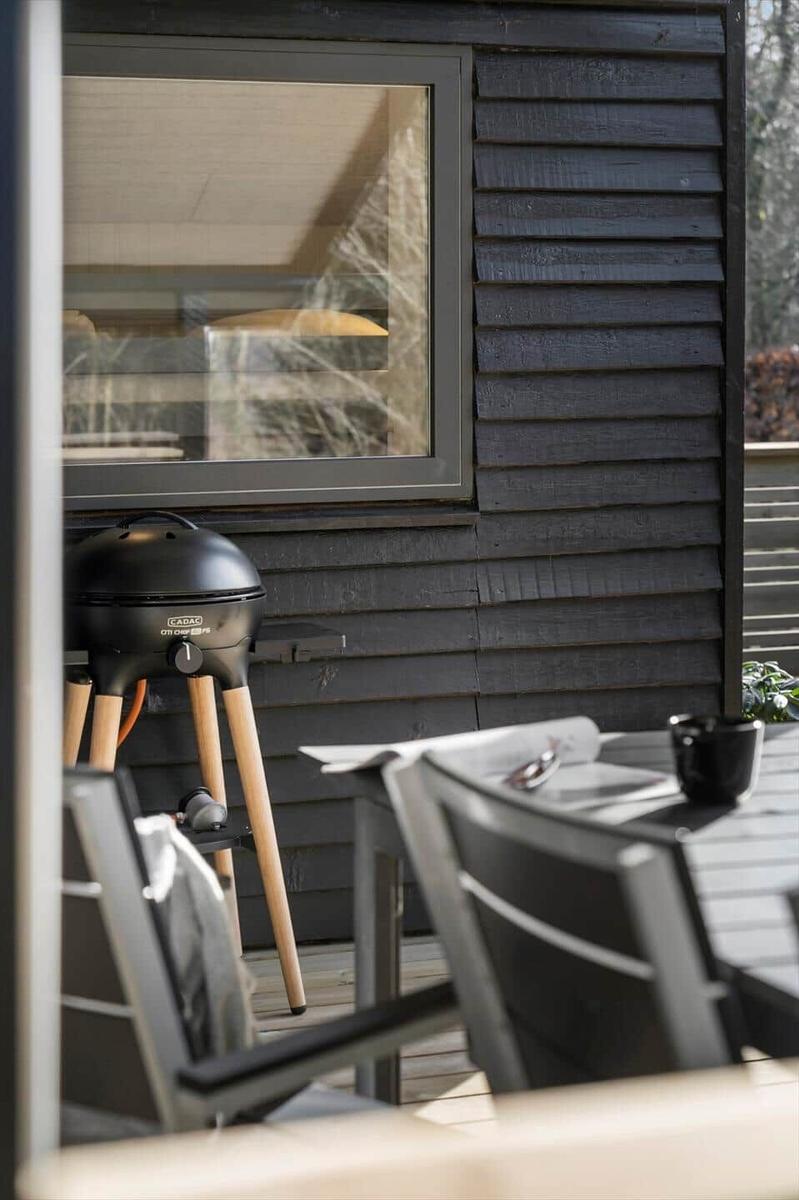 Außenbereich mit Grill, Tisch und Stühlen vor einem Haus mit dunkler Holzfassade.