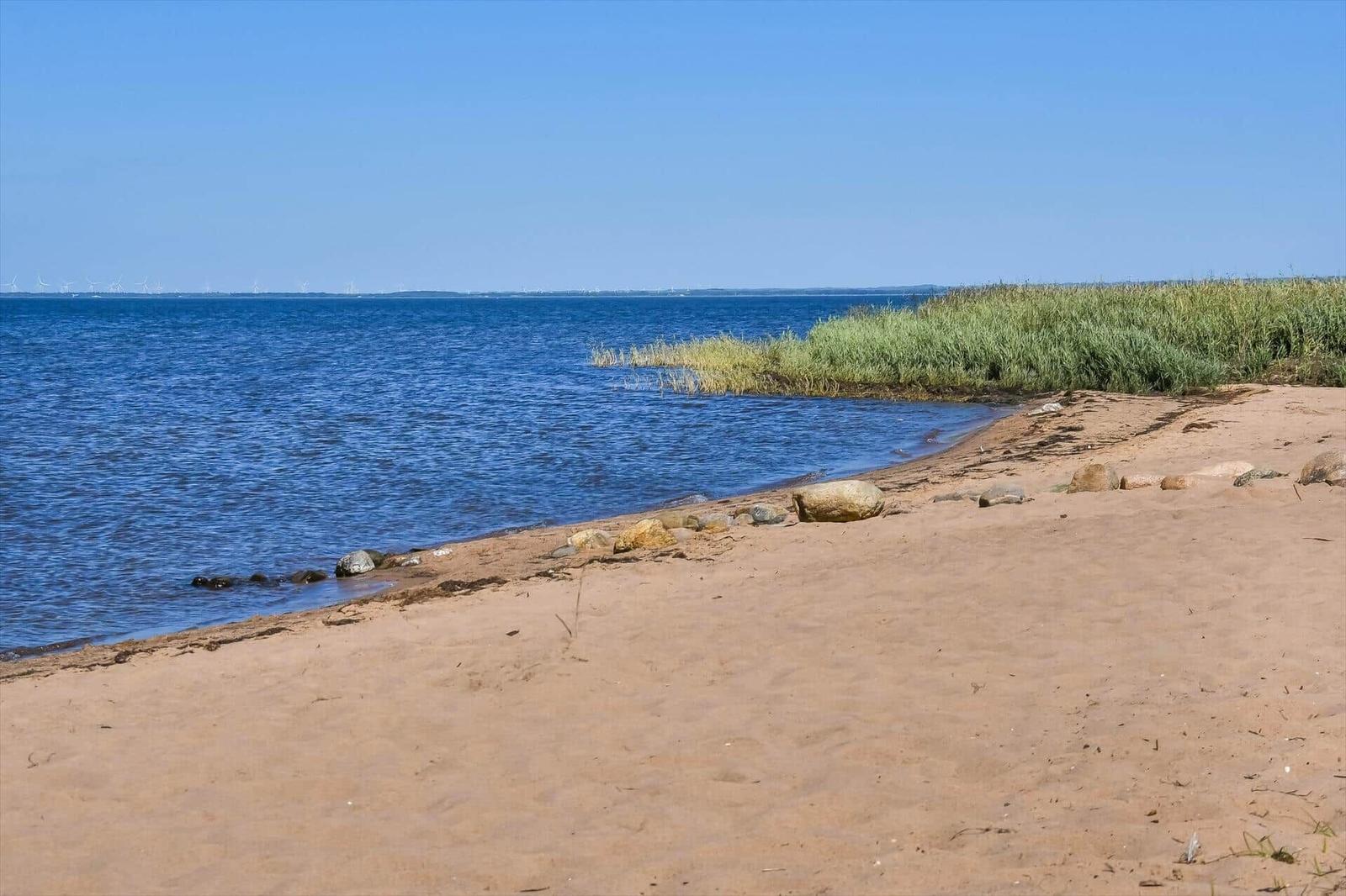 Strand mit Sand, Steinen und Gras am Wasser. Im Hintergrund sind Windräder sichtbar.