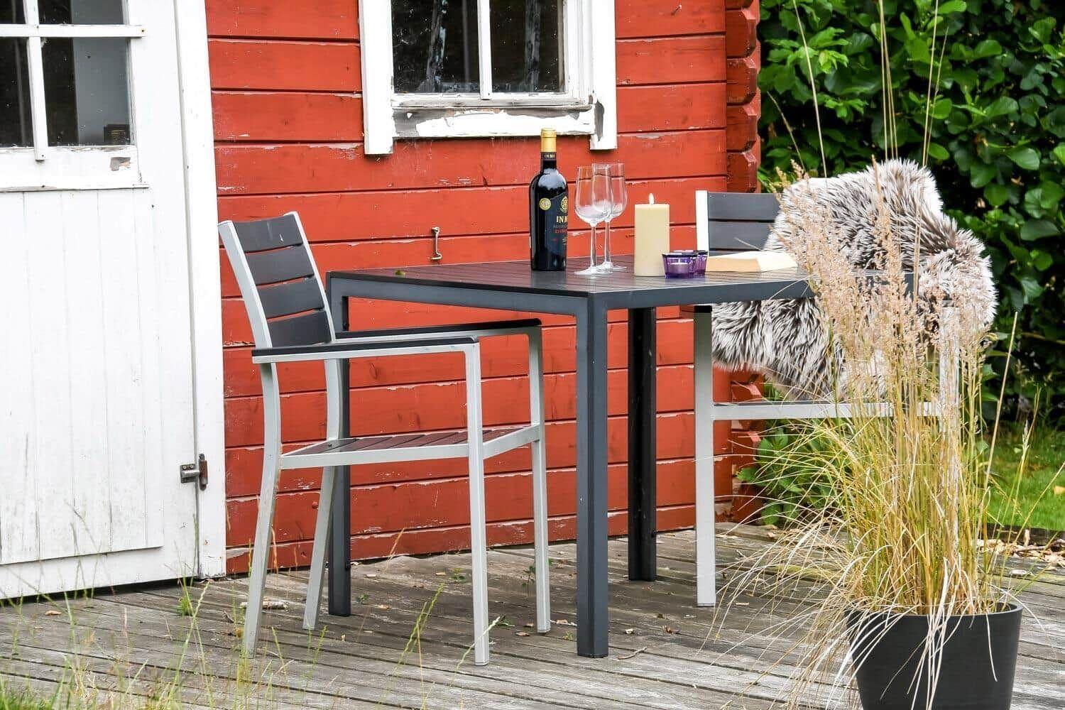 Außentisch mit Stühlen vor rotem Holzhaus, Weinflasche, Gläser und Kerze.
