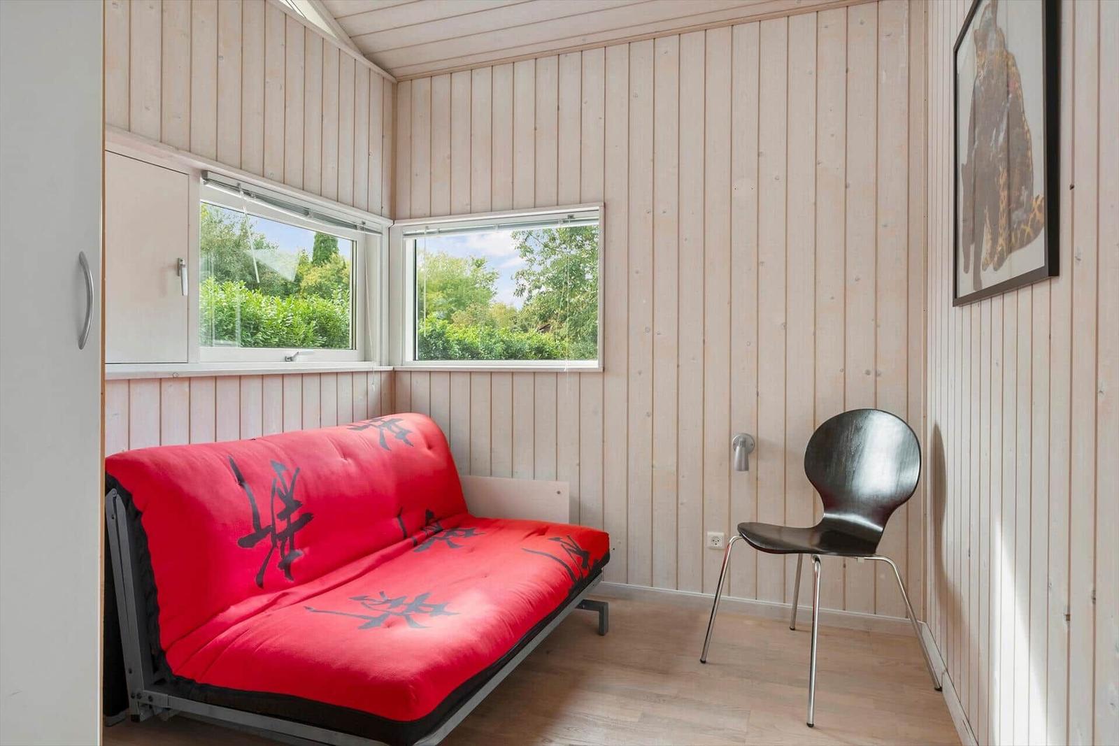 Raum mit rotem Futon, schwarzer Stuhl und Holzwand. Zwei Fenster zeigen grüne Bäume.