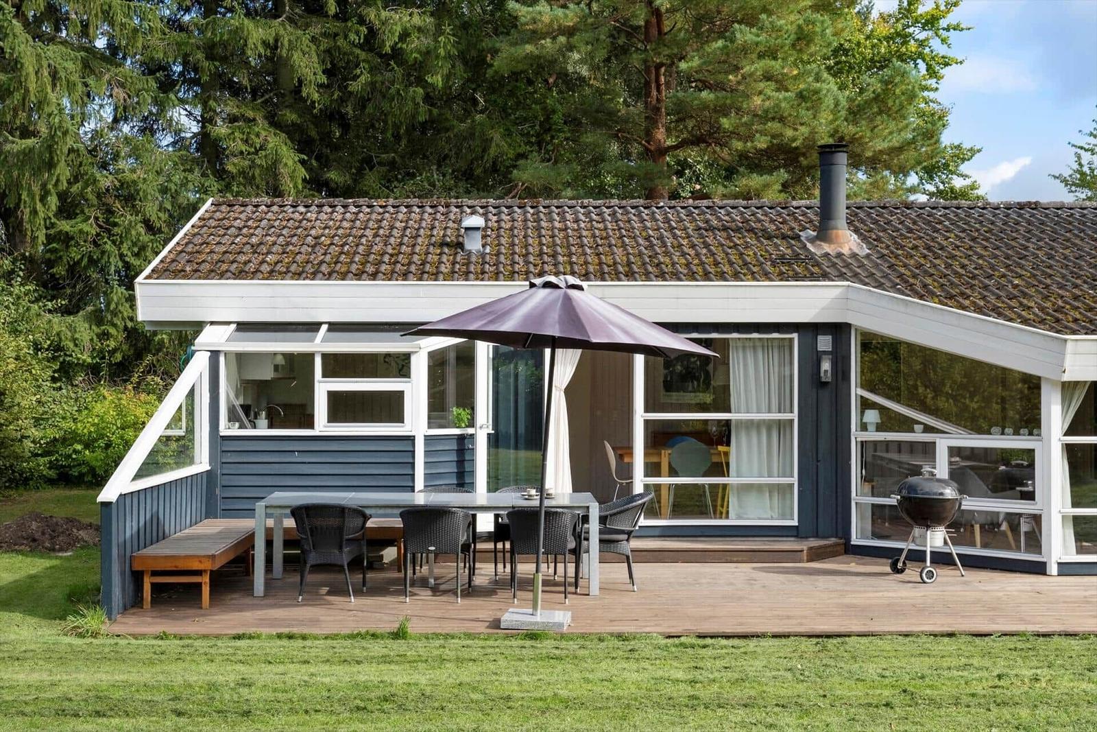 Ein Ferienhaus mit Terrasse, Tisch, Stühlen und Sonnenschirm. Grill und Wald im Hintergrund.