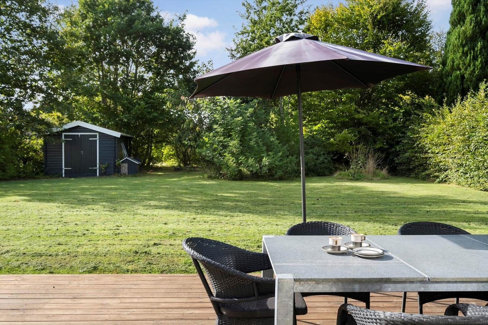 Terrasse mit Tisch, Stühlen und Sonnenschirm. Grünfläche und Holzdecke. Hintergrund mit Gartenhaus und Bäumen.