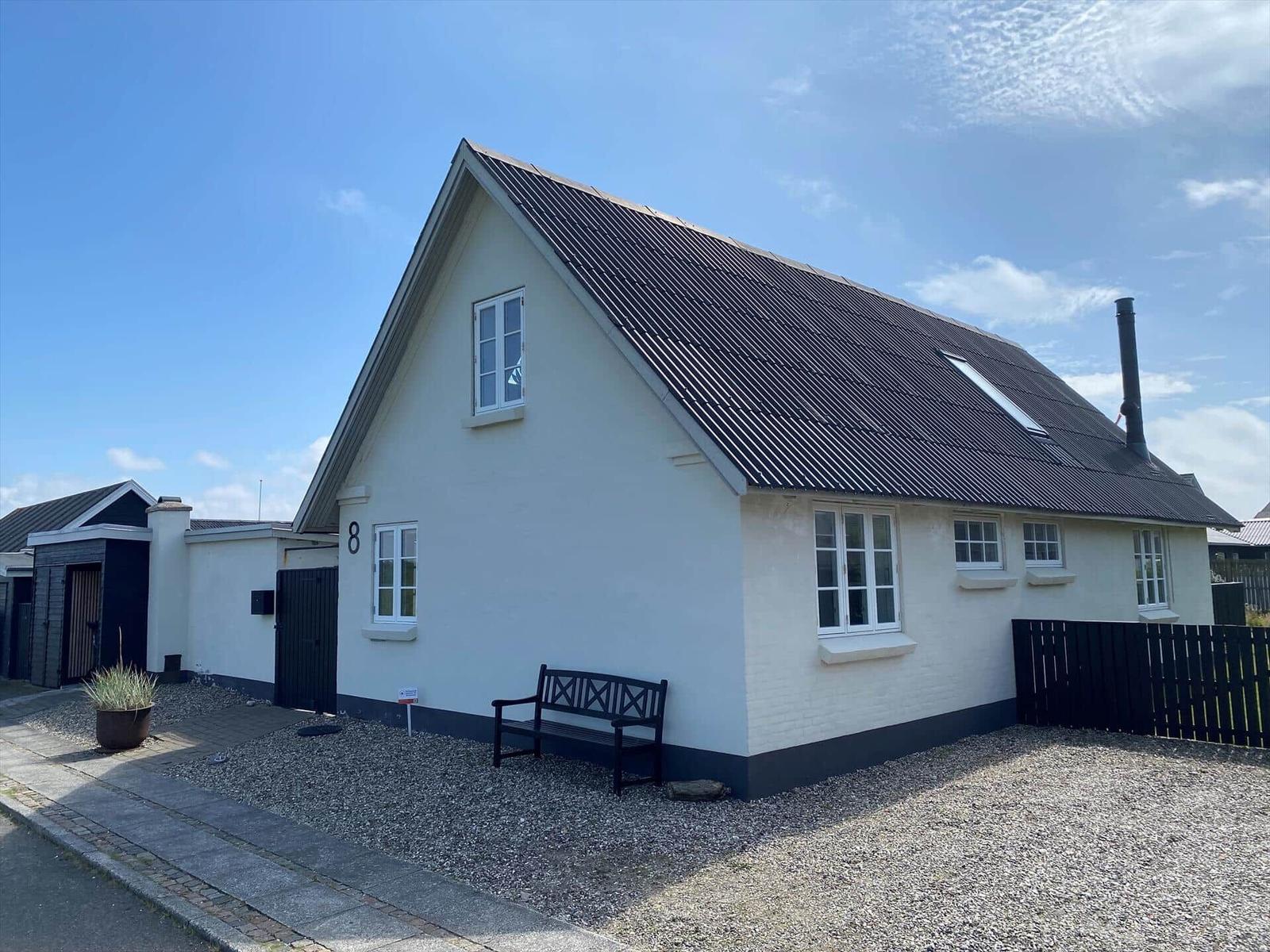 Weißes Haus mit schwarzem Dach und Fenstern. Vorgarten mit Bank und Pflanze.