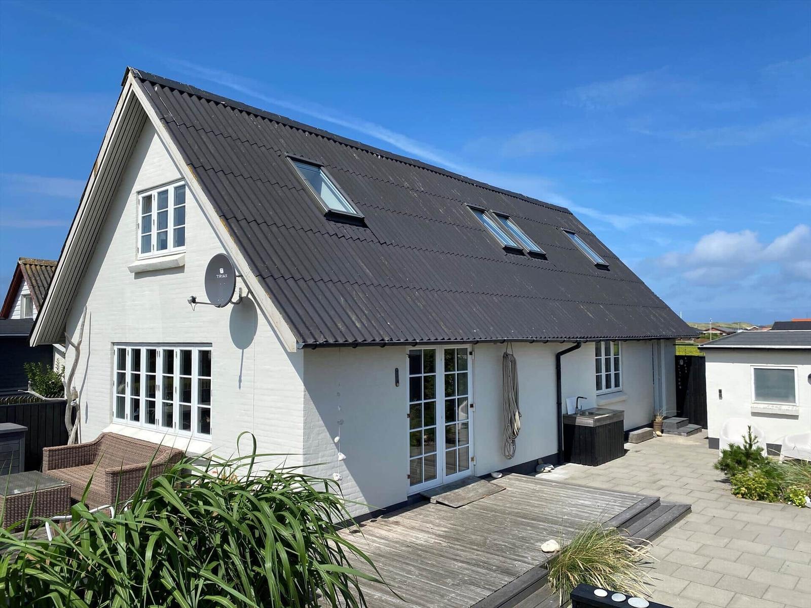 Weißes Haus mit dunklem Dach, Terrasse und Gartenbereich unter blauem Himmel.