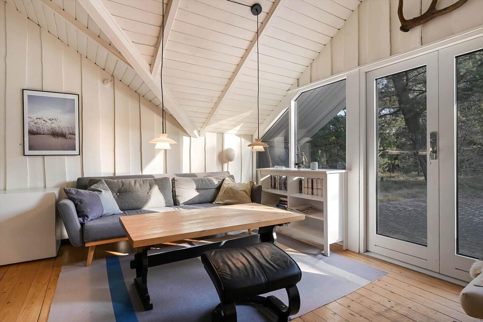 Wohnraum mit Sofa, Tisch und Bücherregal. Holzdecke und große Fenster.