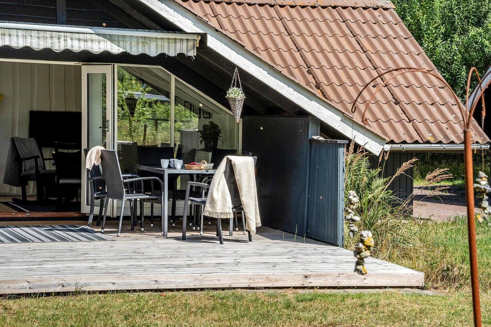 Terrasse mit Tisch und Stühlen unter einem Vordach. Im Hintergrund ist das Haus mit Fenstern zu sehen.