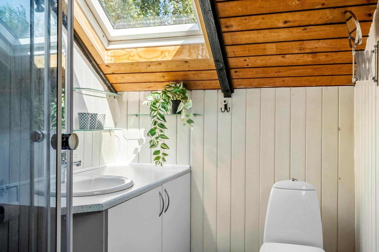 Badezimmer mit Dusche, Waschbecken und Toilette unter Holzdecke mit Dachfenster.
