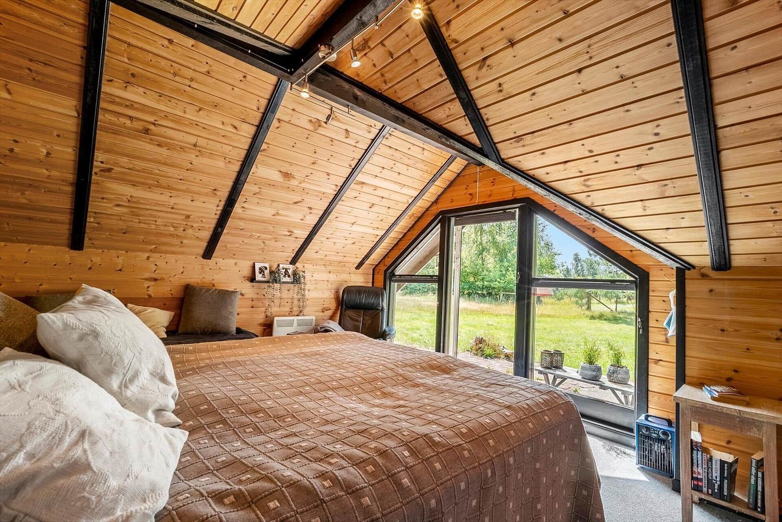 Schlafzimmer mit Holzwand und großem Fenster. Bett, Sessel und Bücherregal sind sichtbar.