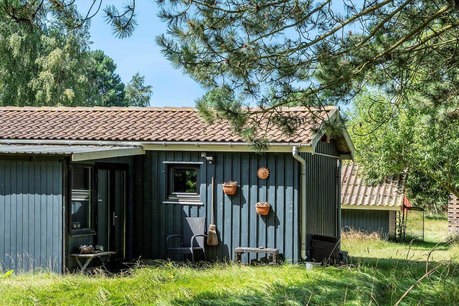 Ein kleines Haus mit grauer Holzfassade und braunem Dach, umgeben von Gras und Bäumen.