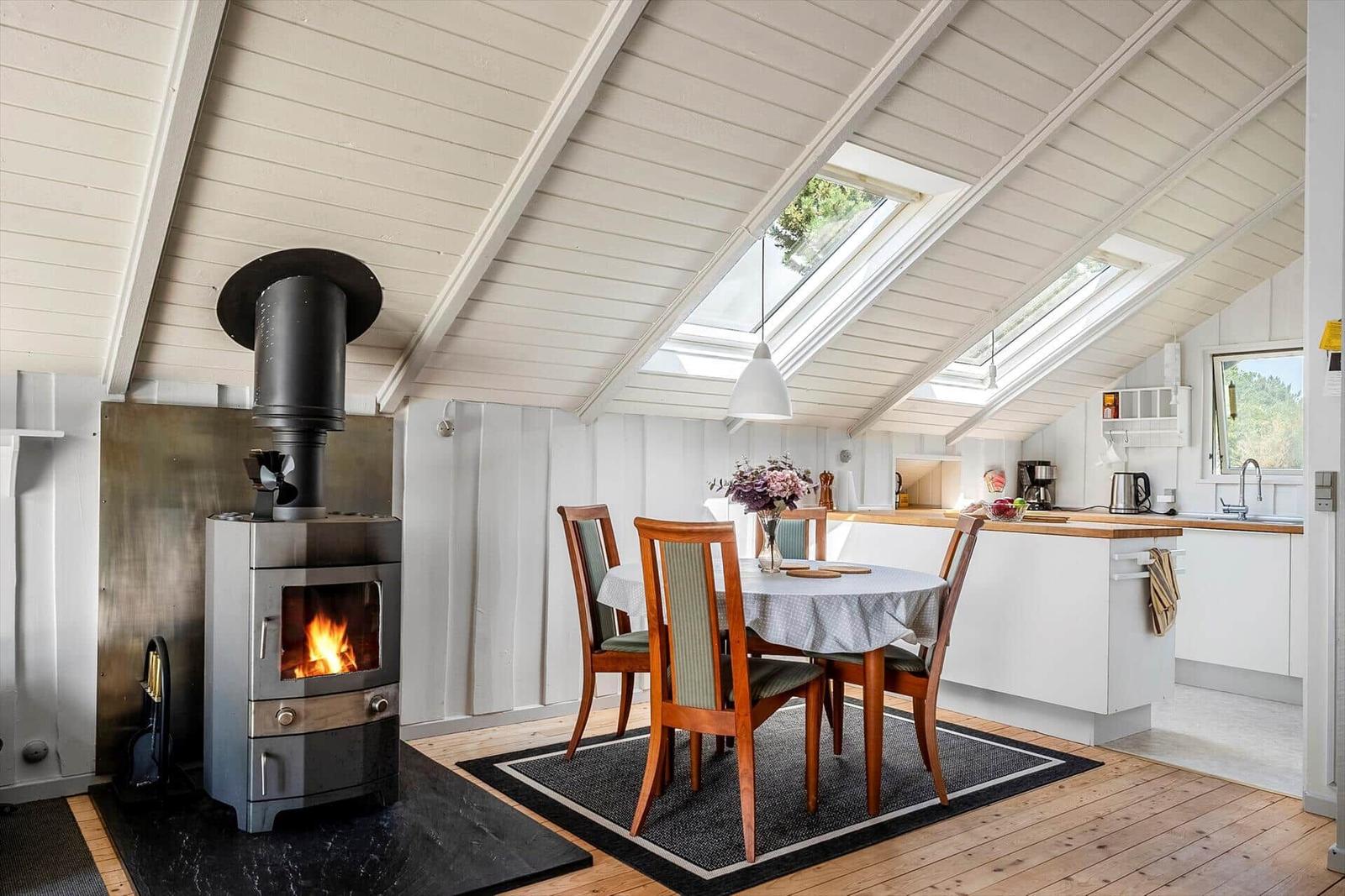 Küche und Essbereich mit Holzboden und Dachfenstern. Holzstühle um Tisch. Kamin mit brennendem Feuer.