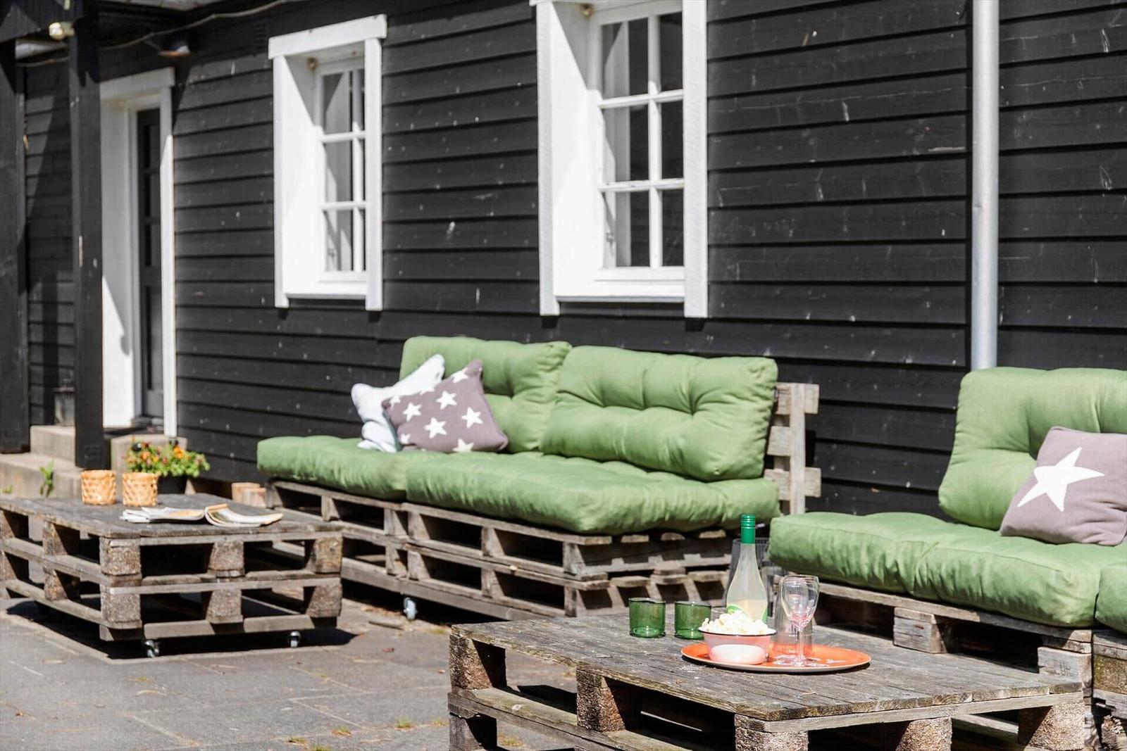 Außenbereich mit grünen Sofas und Holztischen aus Paletten vor schwarzem Haus.