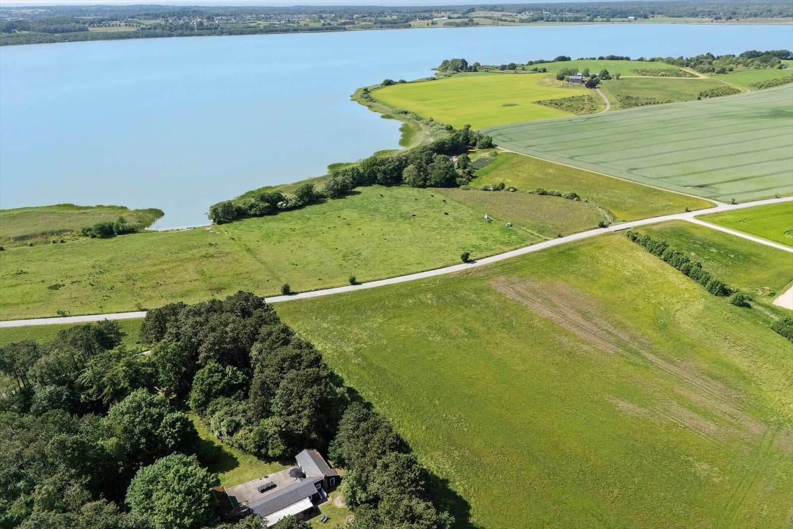 Haus am See mit grünen Feldern und Wald. Luftbild zeigt ruhige Landschaft.