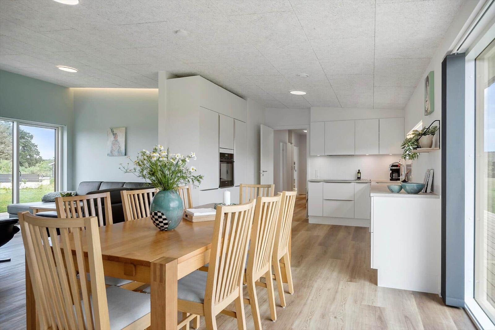 Essbereich mit Holztisch und Stühlen. Küche mit weißen Schränken. Wohnbereich mit Sofa und Fenster.