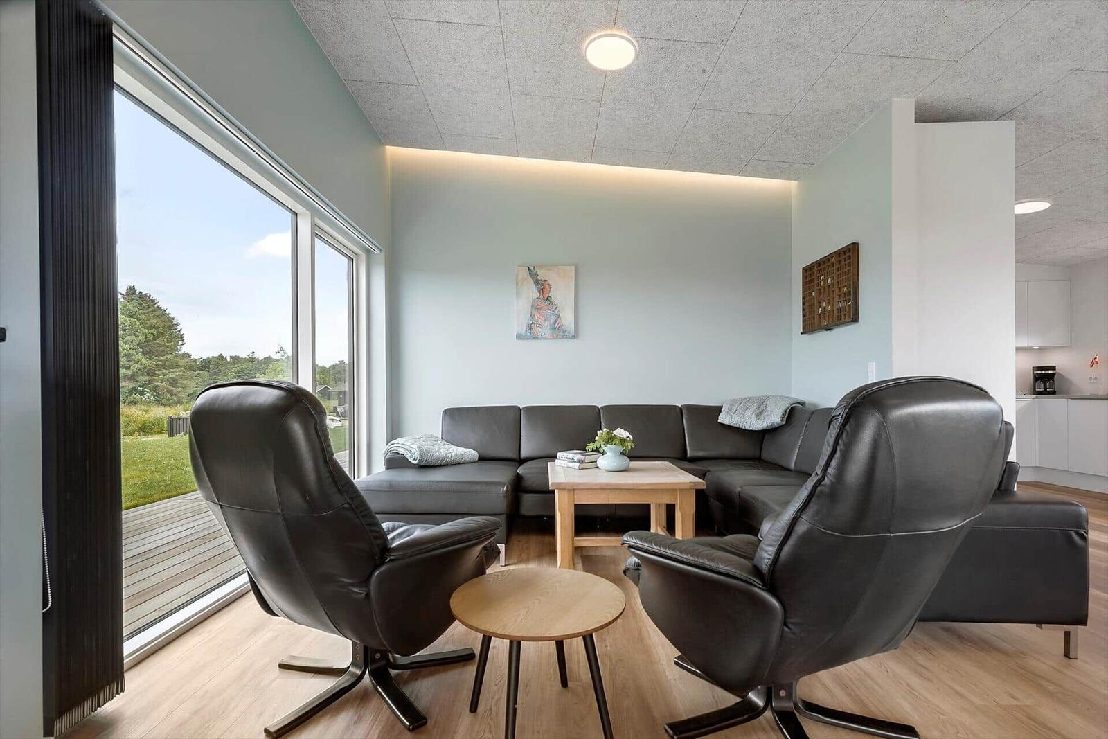 Wohnzimmer mit Sofa, zwei Sesseln und großem Fenster zur Außenterrasse.