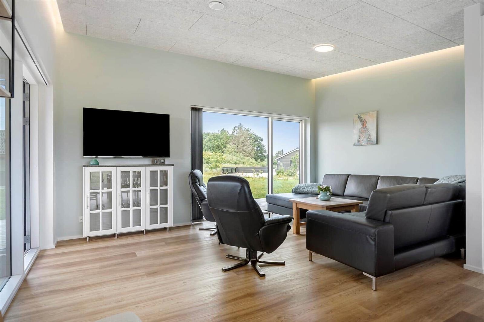 Wohnzimmer mit Sofa, Sessel und Fernseher. Große Fenster mit Blick auf Grünfläche.