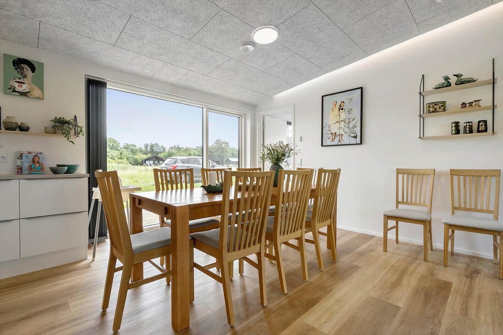 Esszimmer mit Holztisch, Stühlen und großem Fenster. Wandregale und Bilder an den Wänden.
