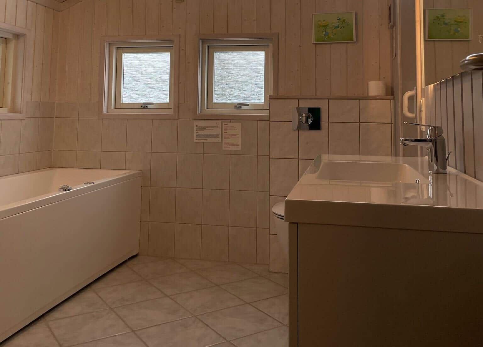 Badezimmer mit Badewanne, Waschbecken und Toilette. Holzwände und Fenster mit Blick auf Wasser.