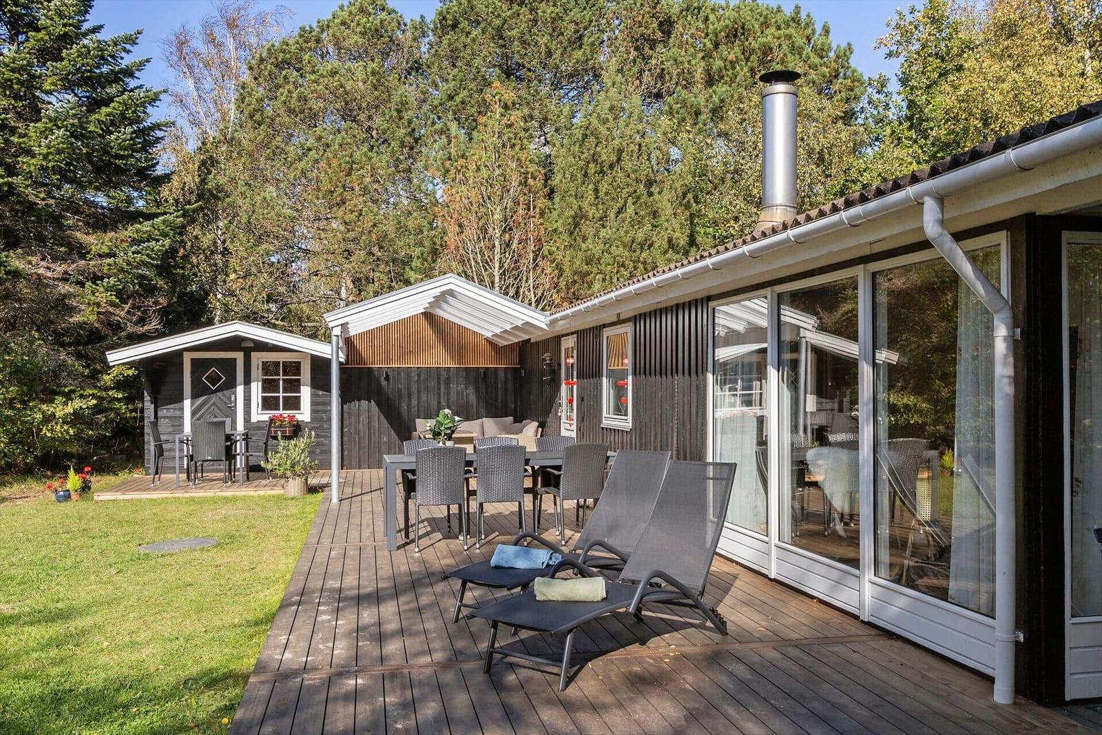 Terrasse mit Liegestühlen, Tisch und Sitzgelegenheiten. Hintergrund: Wald und kleines Haus.