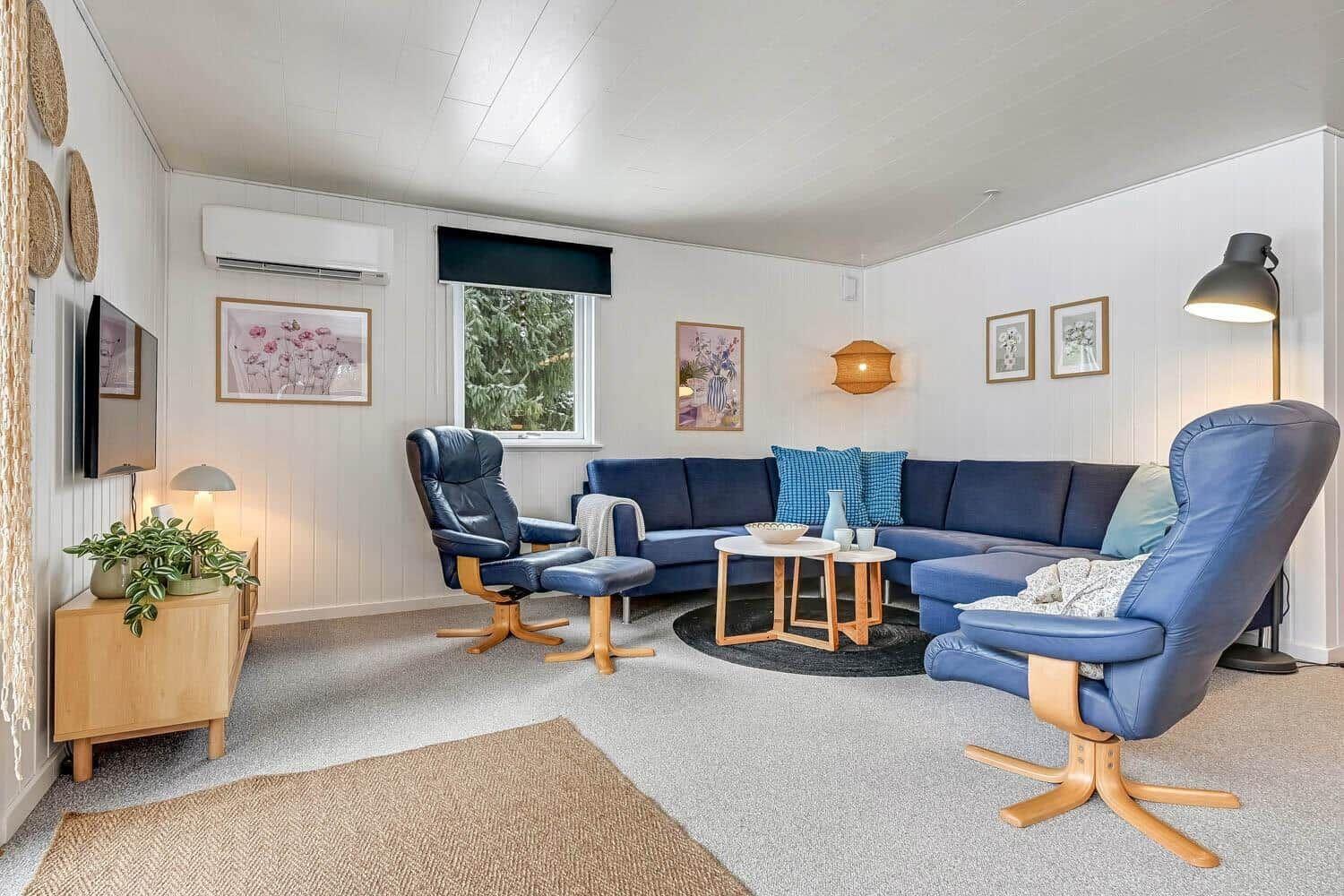 Wohnzimmer mit blauem Sofa, zwei Sesseln und Holztisch. Wandbilder und Pflanzen.