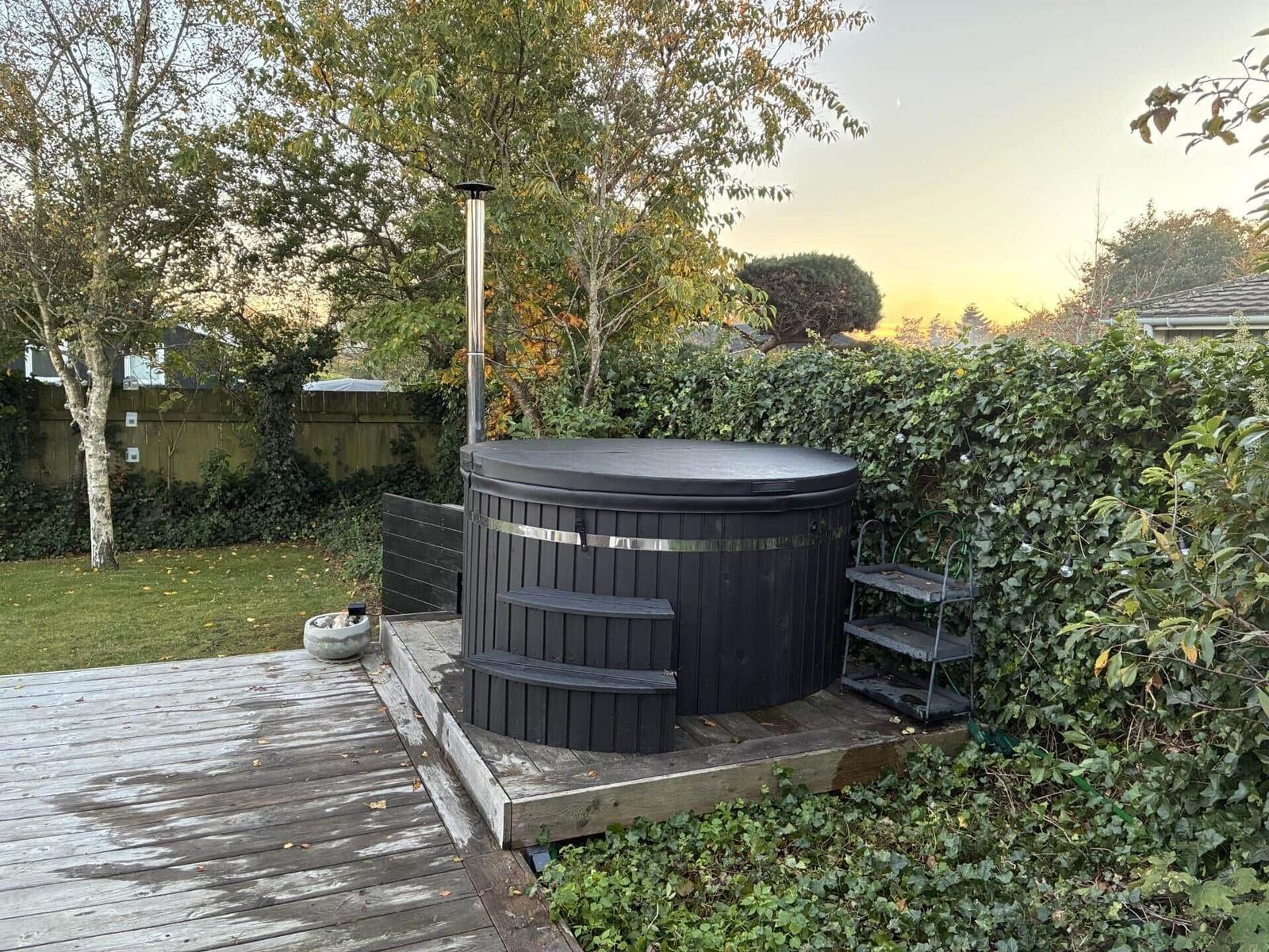 Schwarzer Whirlpool auf Holzterrasse mit Garten und Bäumen im Hintergrund.