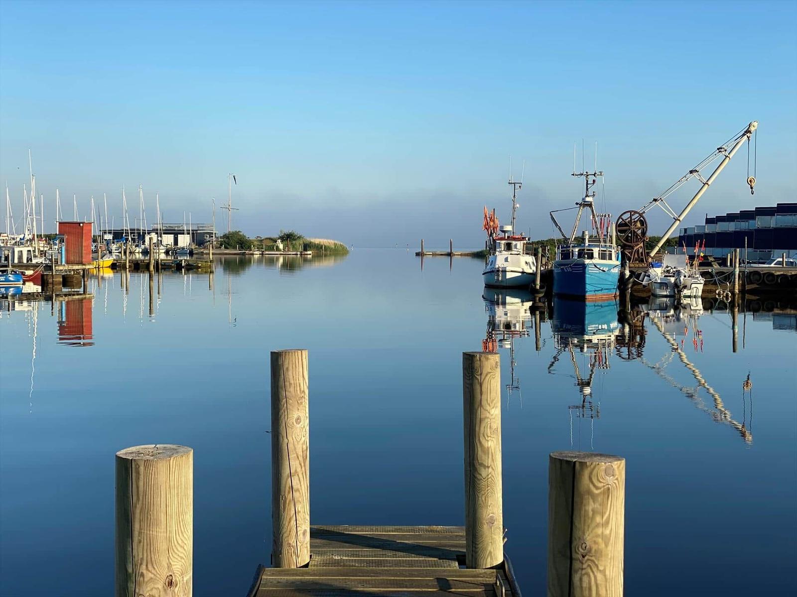 Hafen mit Booten und Steg im Vordergrund. Spiegelglatte Wasseroberfläche reflektiert die Schiffe.