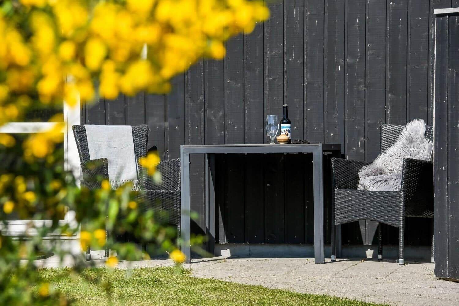Außenbereich mit Tisch und zwei Stühlen vor schwarzem Holzhaus. Gelbe Blumen im Vordergrund.