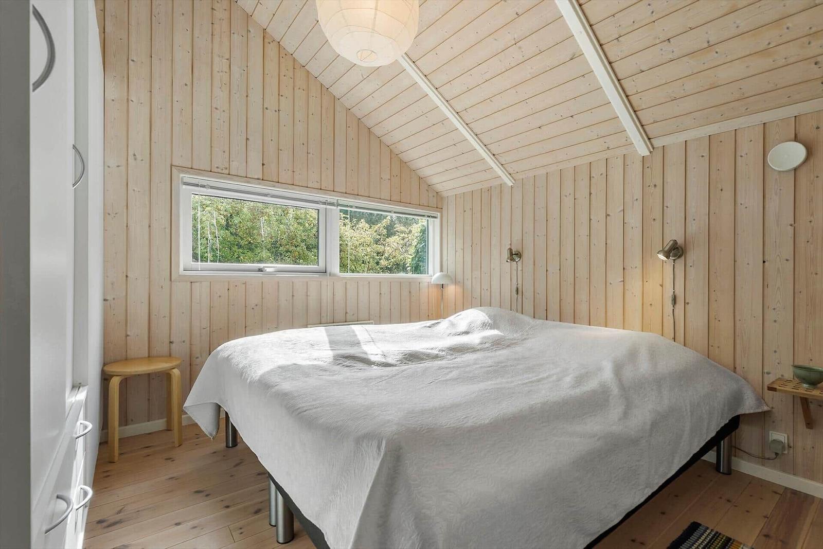 Bedroom with wooden walls and sloped ceiling. Bed, nightstand, stool, and wardrobe.