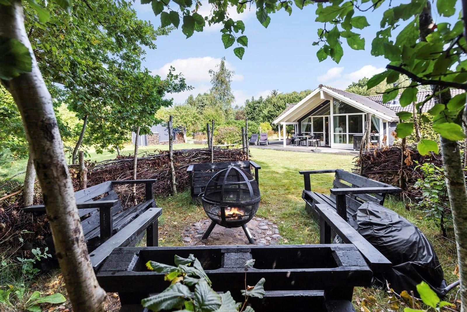 Behind a wooden fence is a fire pit with burning wood. In the background, a house with a terrace is visible.