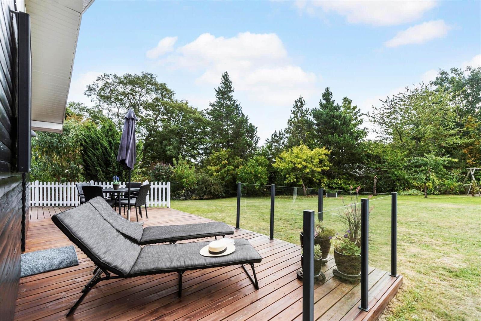 Terrasse mit Liegestühlen, Tisch und Gartenzaun. Blick auf grünen Rasen und Bäume.