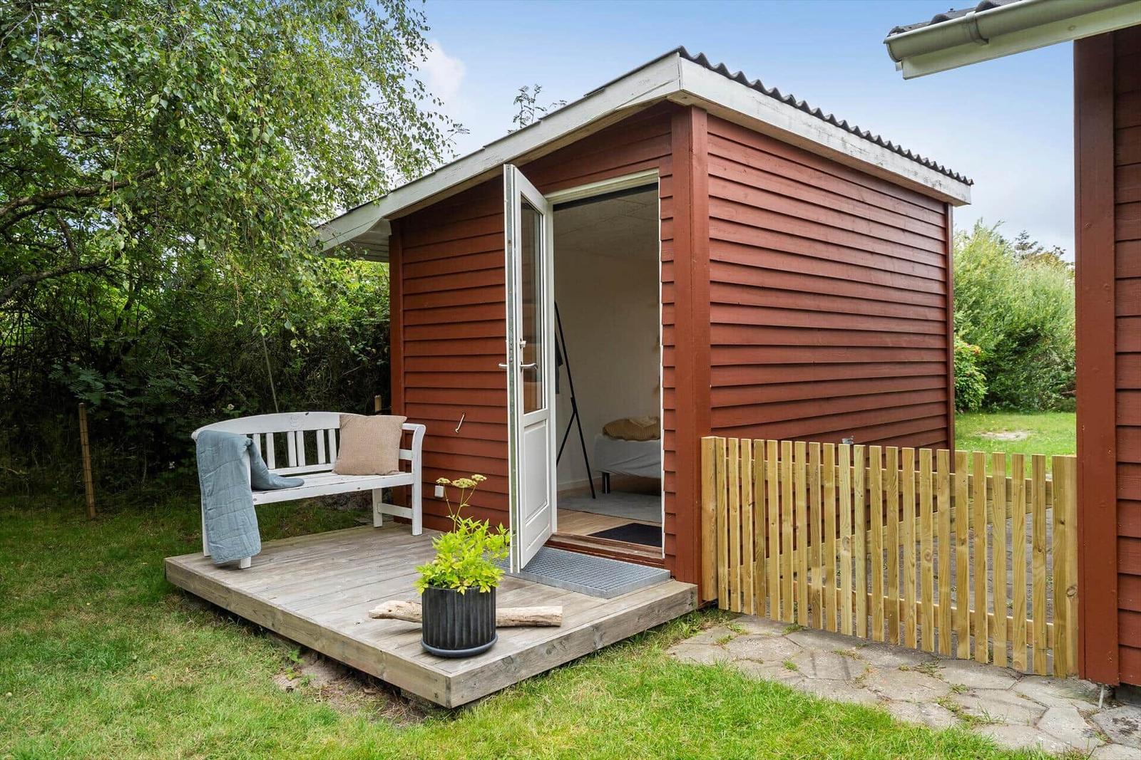 Rotes Holzhaus mit offener Tür, weißer Bank und grünem Garten.