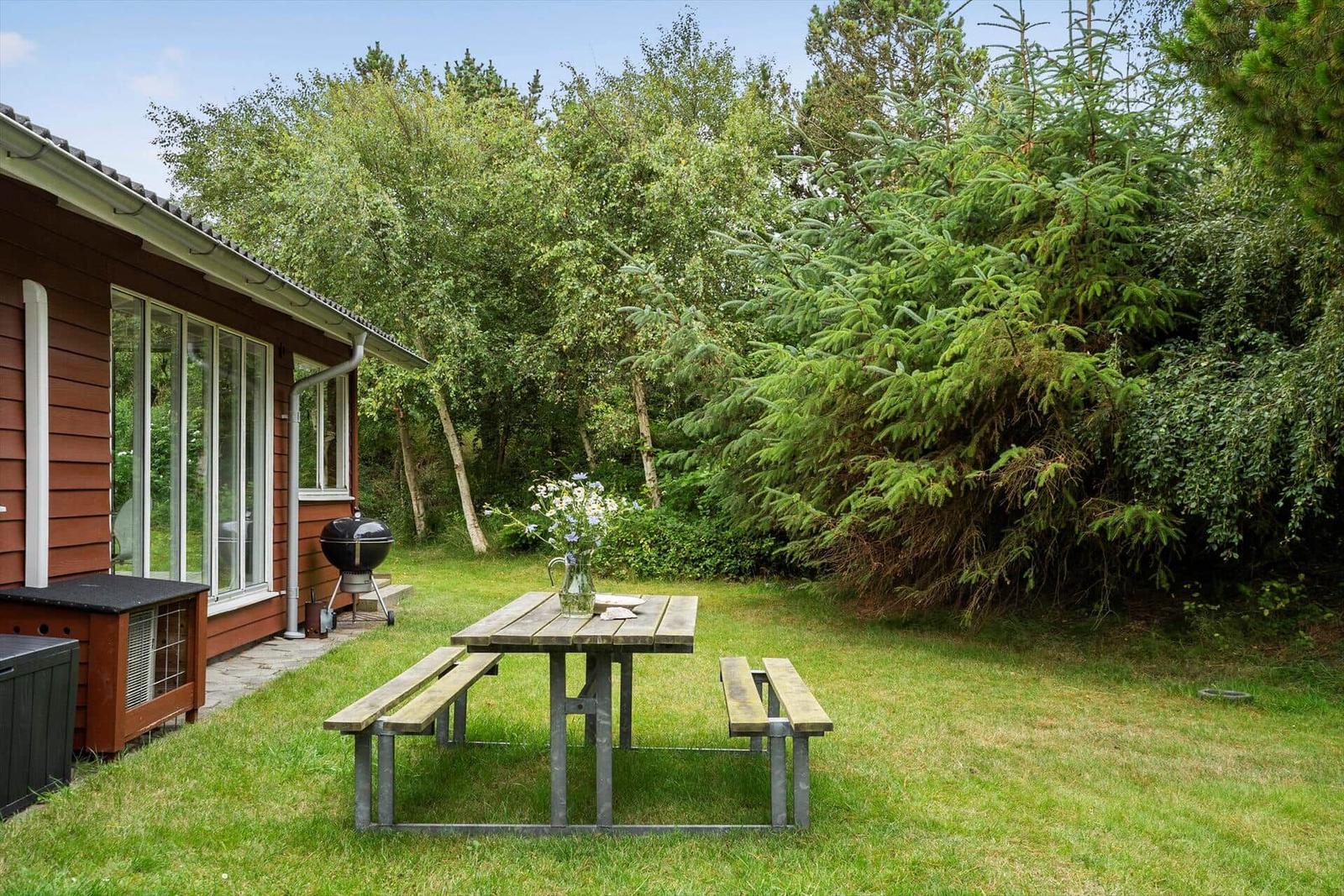 Hinter dem Haus liegt ein Garten mit Tisch, Bänken und Grill. Umgeben von Bäumen und Sträuchern.