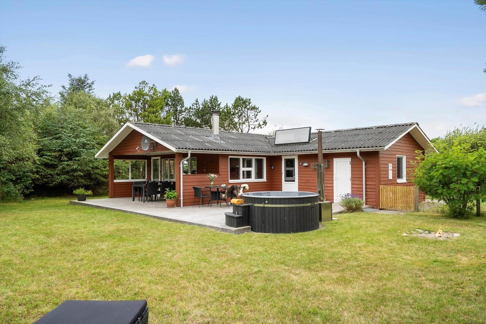 Rotes Ferienhaus mit Terrasse, Hot tub und Garten. Hintergrund: Bäume und klarer Himmel.