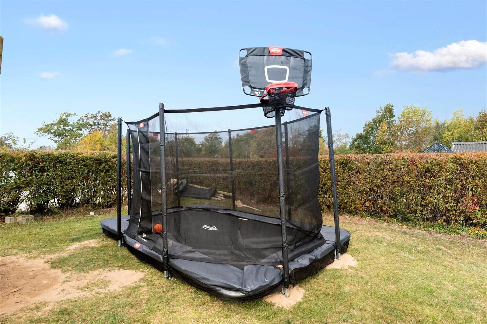 Trampolin mit Korb im Garten, umgeben von Hecke und Gras.