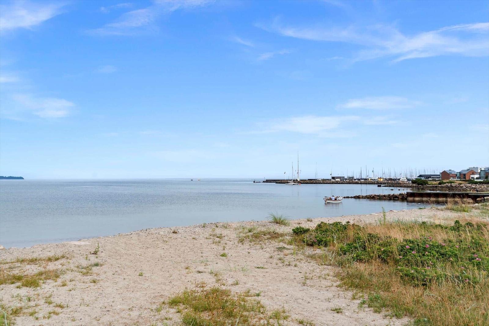 Strand mit Blick auf Hafen und Segelboote unter blauem Himmel.