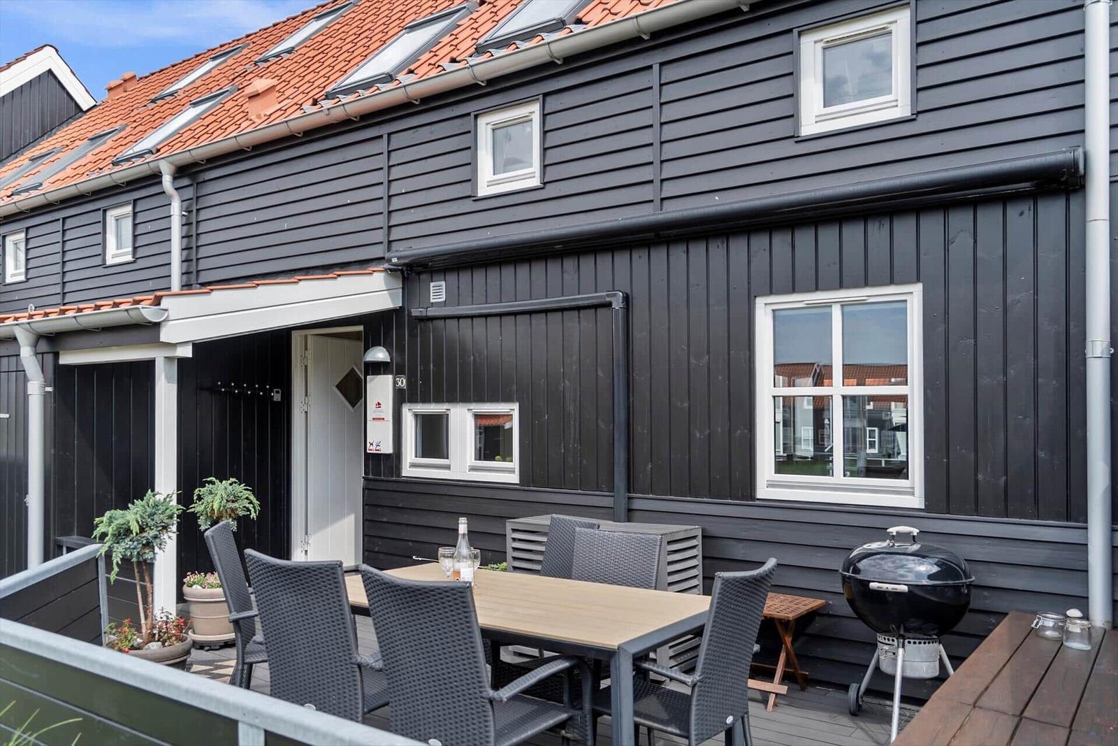 Außenbereich mit Tisch, Stühlen und Grill vor einem Haus mit dunkler Fassade und rotem Dach.