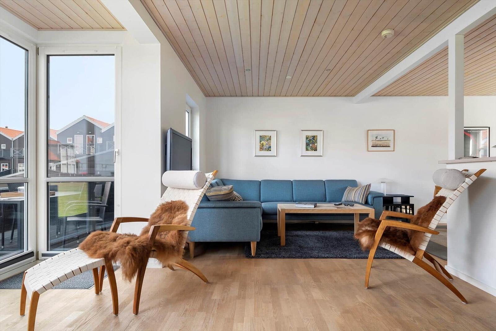 Ein Wohnzimmer mit blauem Sofa, zwei Holzstühlen mit Fell, Holzboden und großen Fenstern.