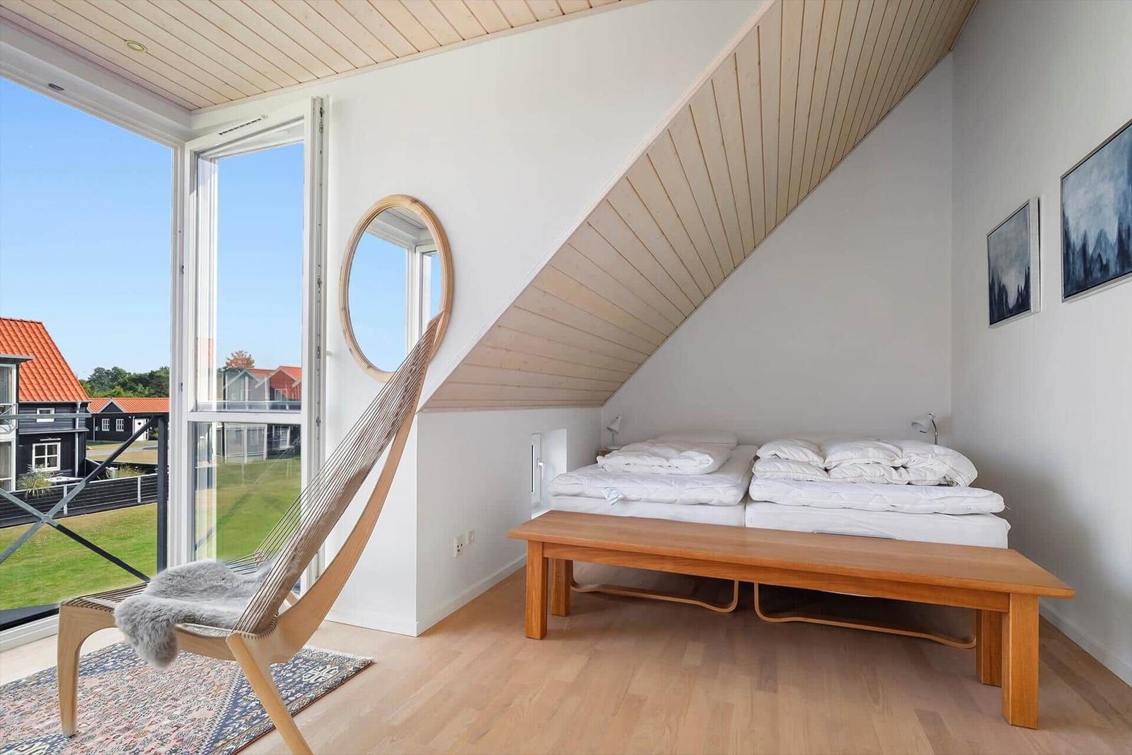 Schlafzimmer mit Dachschräge, Bett, Holzbank und großem Fenster mit Blick auf Garten.