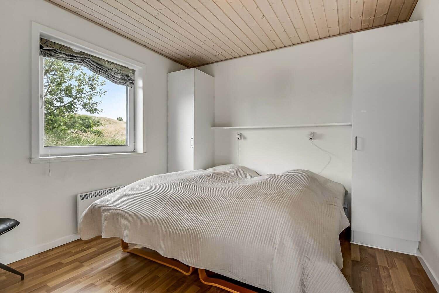 Schlafzimmer mit Bett, Fenster und weißen Schränken. Holzdecke und Parkettboden.