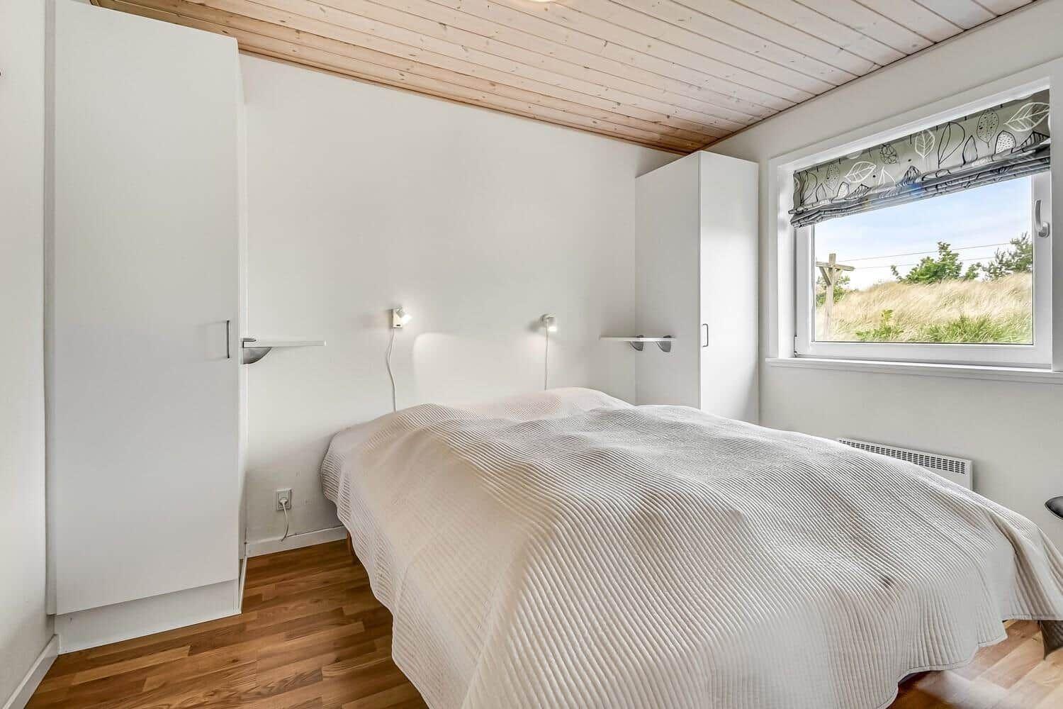 Schlafzimmer mit Bett, weißen Schränken und Holzdecke. Fenster mit Blick auf Gras.