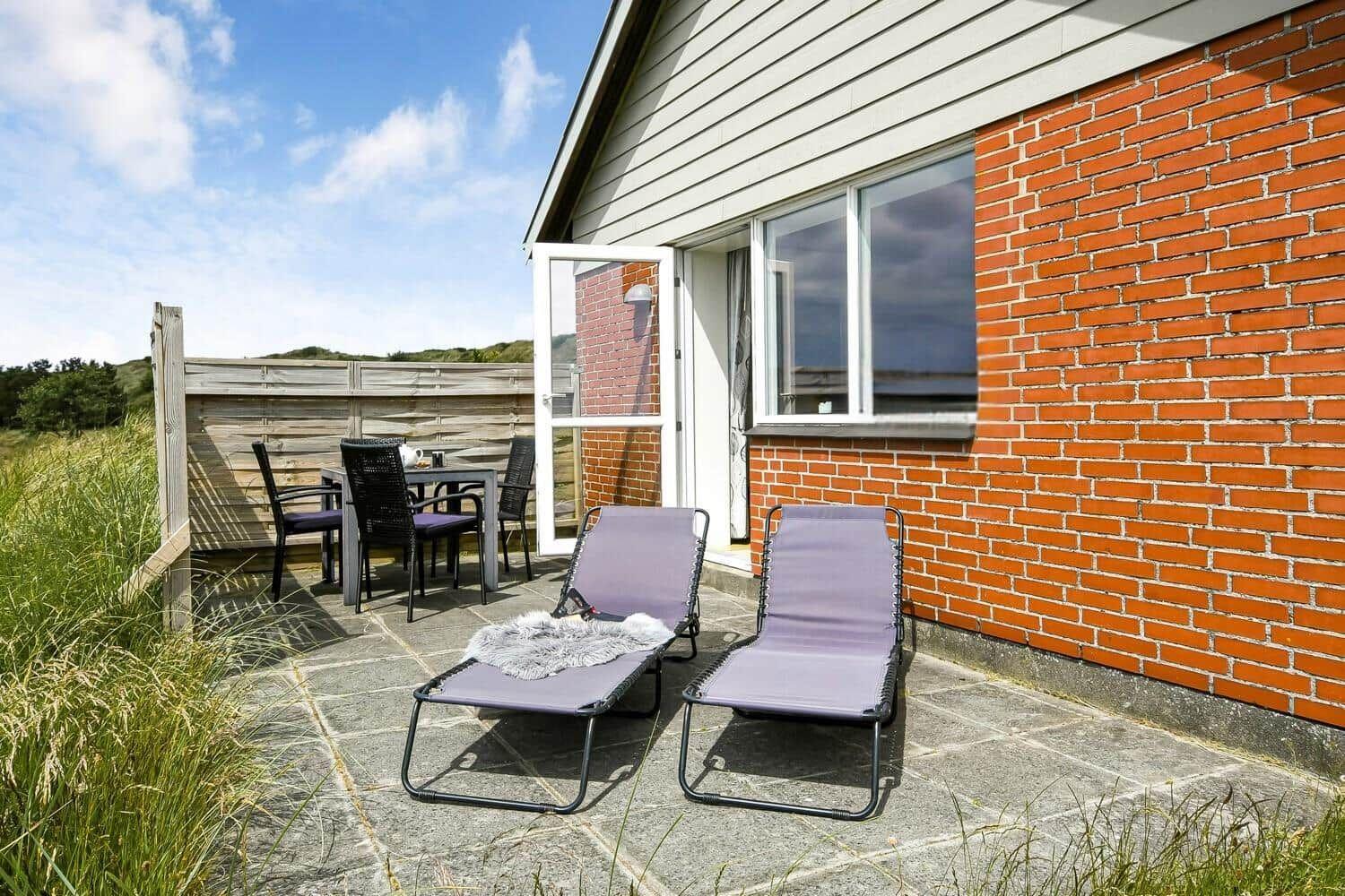 Terrasse mit Liegestühlen und Tisch. Haus mit Backsteinwand und offener Tür.