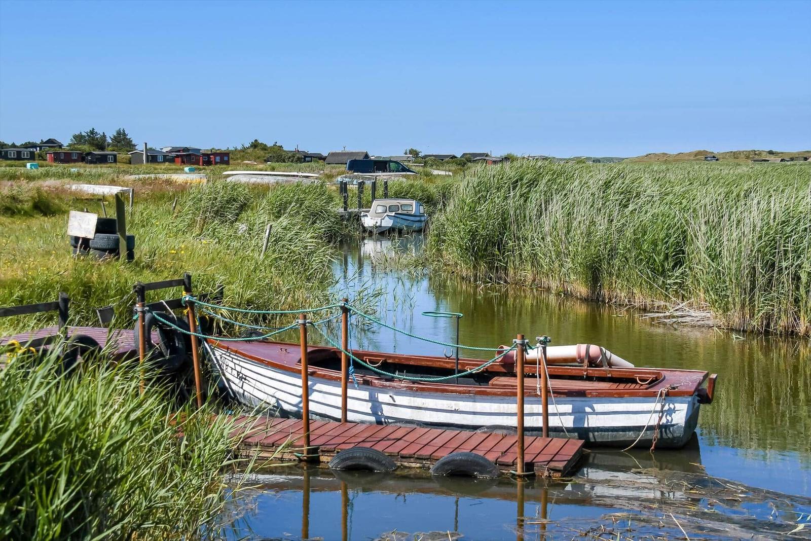 Ein hölzerner Steg führt zu einem Boot im Wasser. Hochgras und Häuser sind im Hintergrund sichtbar.